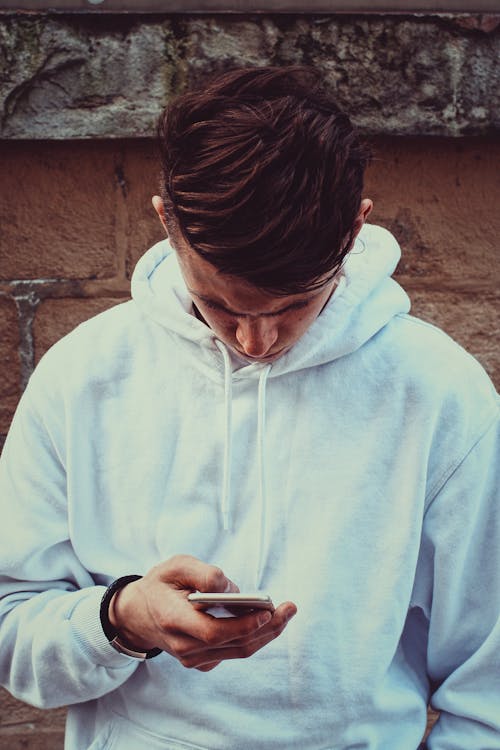 Free Photo of Man in White Hoodie Standing Near Brown Wall While Using His Smartphone Stock Photo