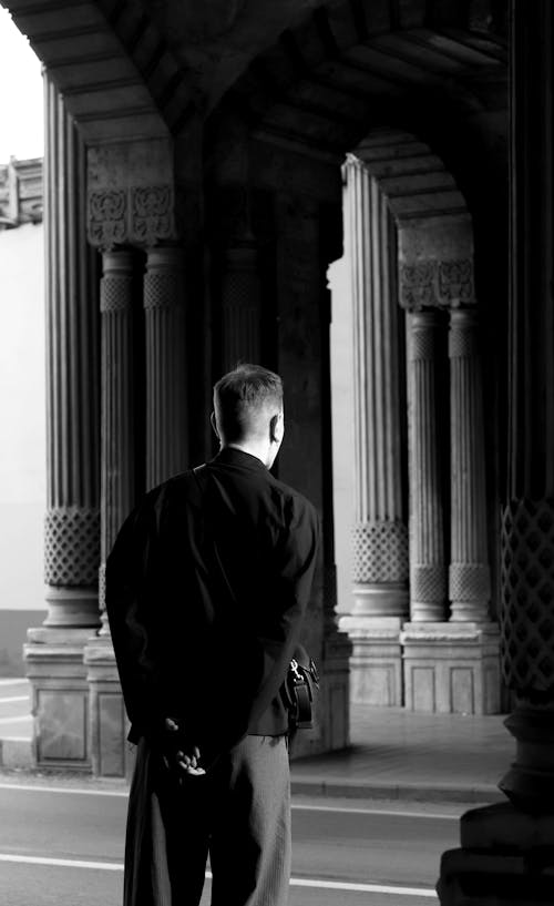 Black and White Photo of a Man Standing under an Archway with Columns in City 