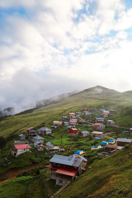 A picturesque mountain village with scattered wooden houses, surrounded by lush green hills and a cloudy sky.