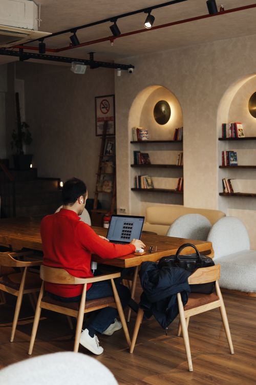 Free A man in a red sweater working on a laptop in a stylish café with wooden furniture and bookshelves. Stock Photo
