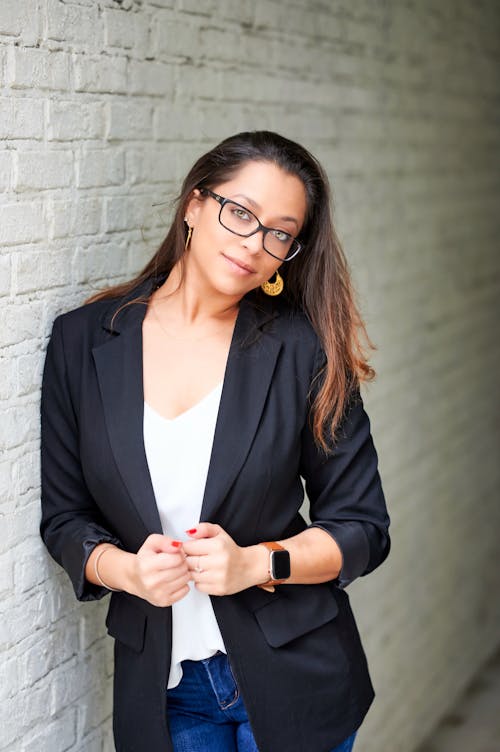 A woman in glasses and a black blazer leaning against a brick wall