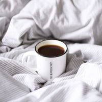 Cup of coffee resting on rumpled white bed linen, photo by Enisa Halidi on Pexels