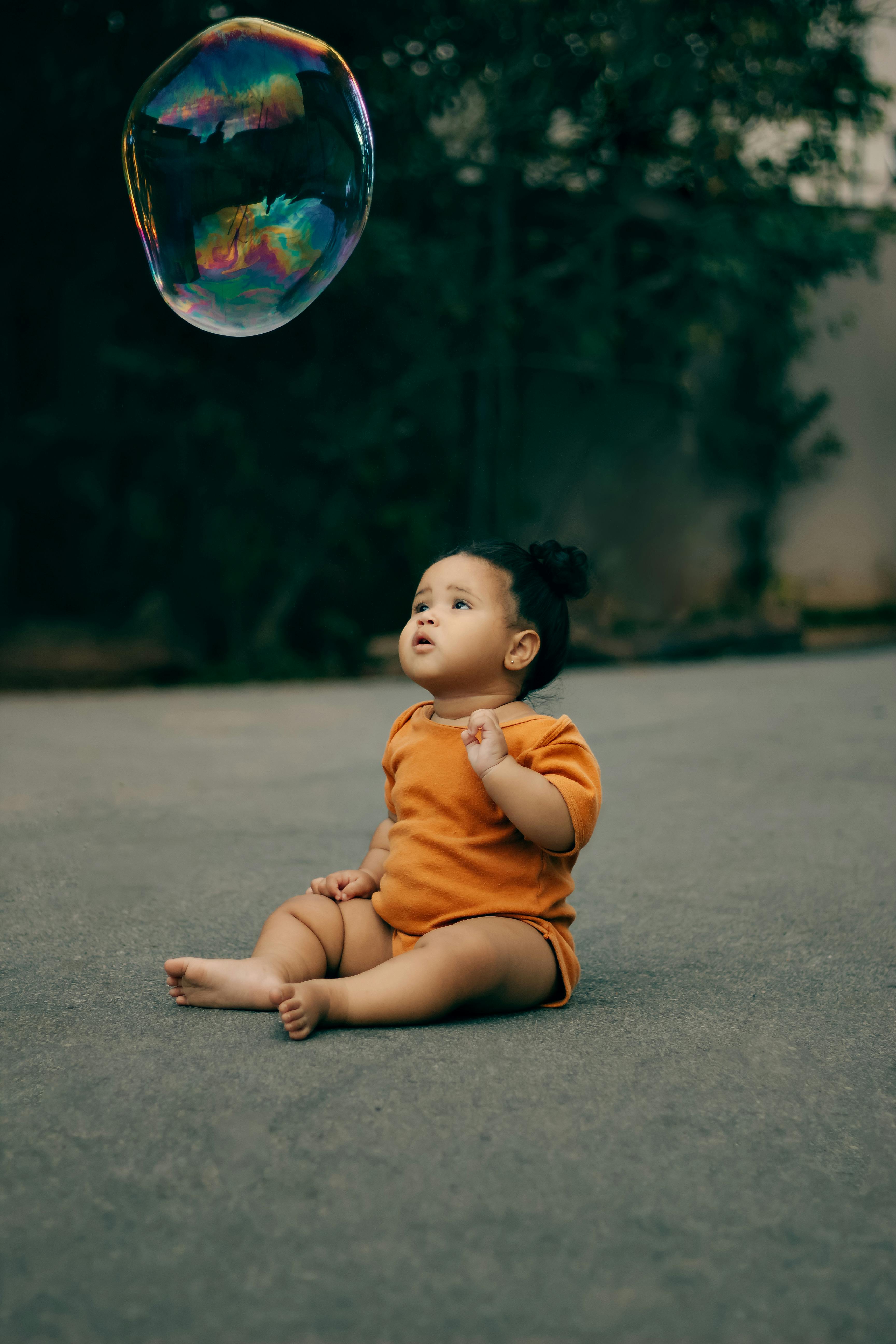 Cute baby in orange clothing sits outdoors, gazing curiously at a floating bubble.