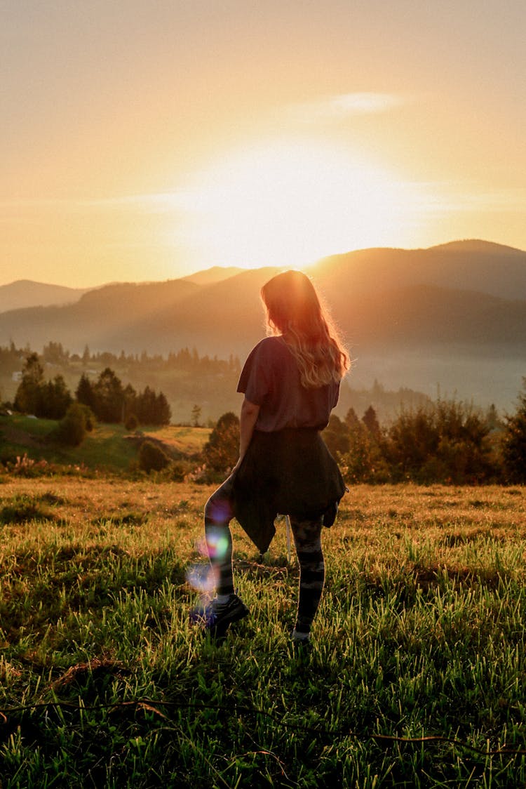 Silhouette Of A Young Woman Standing On The Grassland With The View Of The Mountains In The Sunset