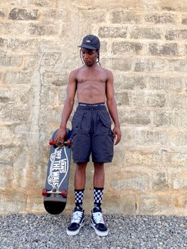 African adult male skater standing shirtless against brick wall holding a skateboard.