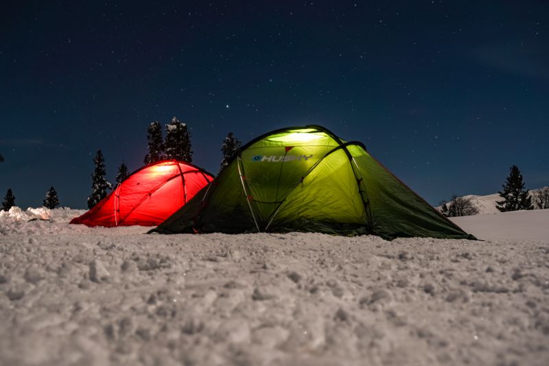 Two tents illuminated at night, set on a snowy landscape beneath a clear starry sky.