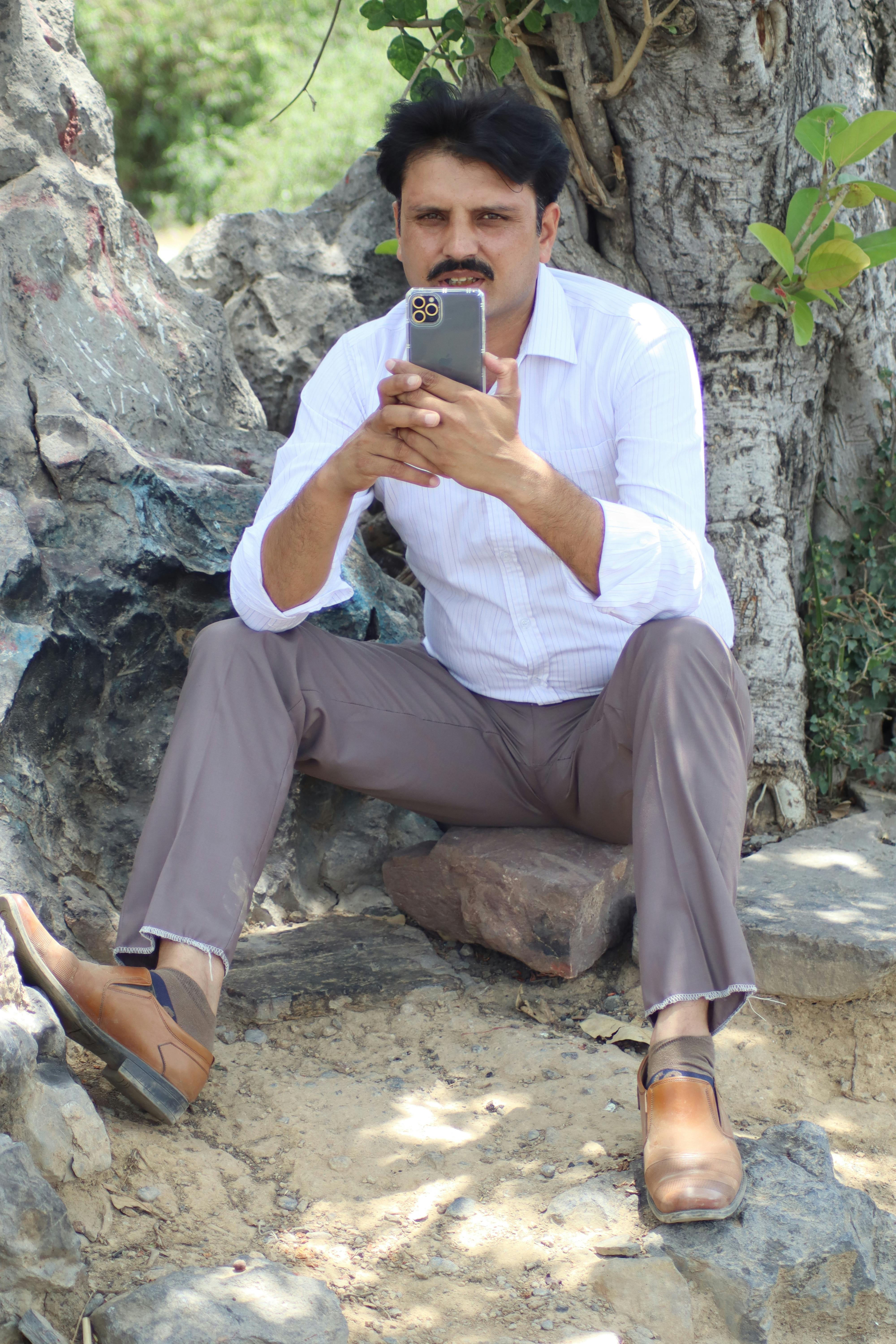 Adult male sitting on rocks, using a smartphone in a natural setting.