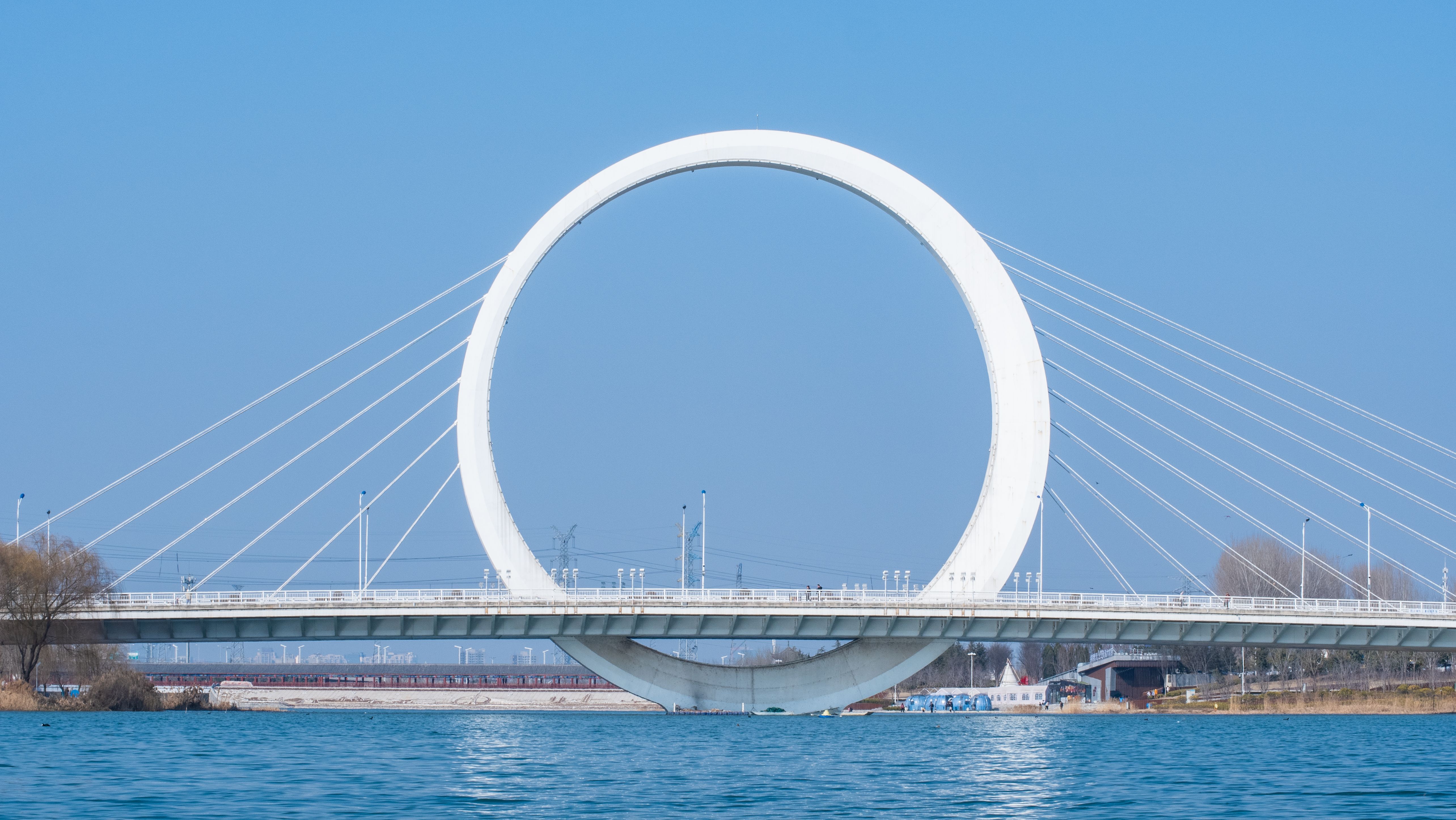 The iconic Infinity Ring Bridge in Zhengzhou, China, spanning over a tranquil river under a clear sky.