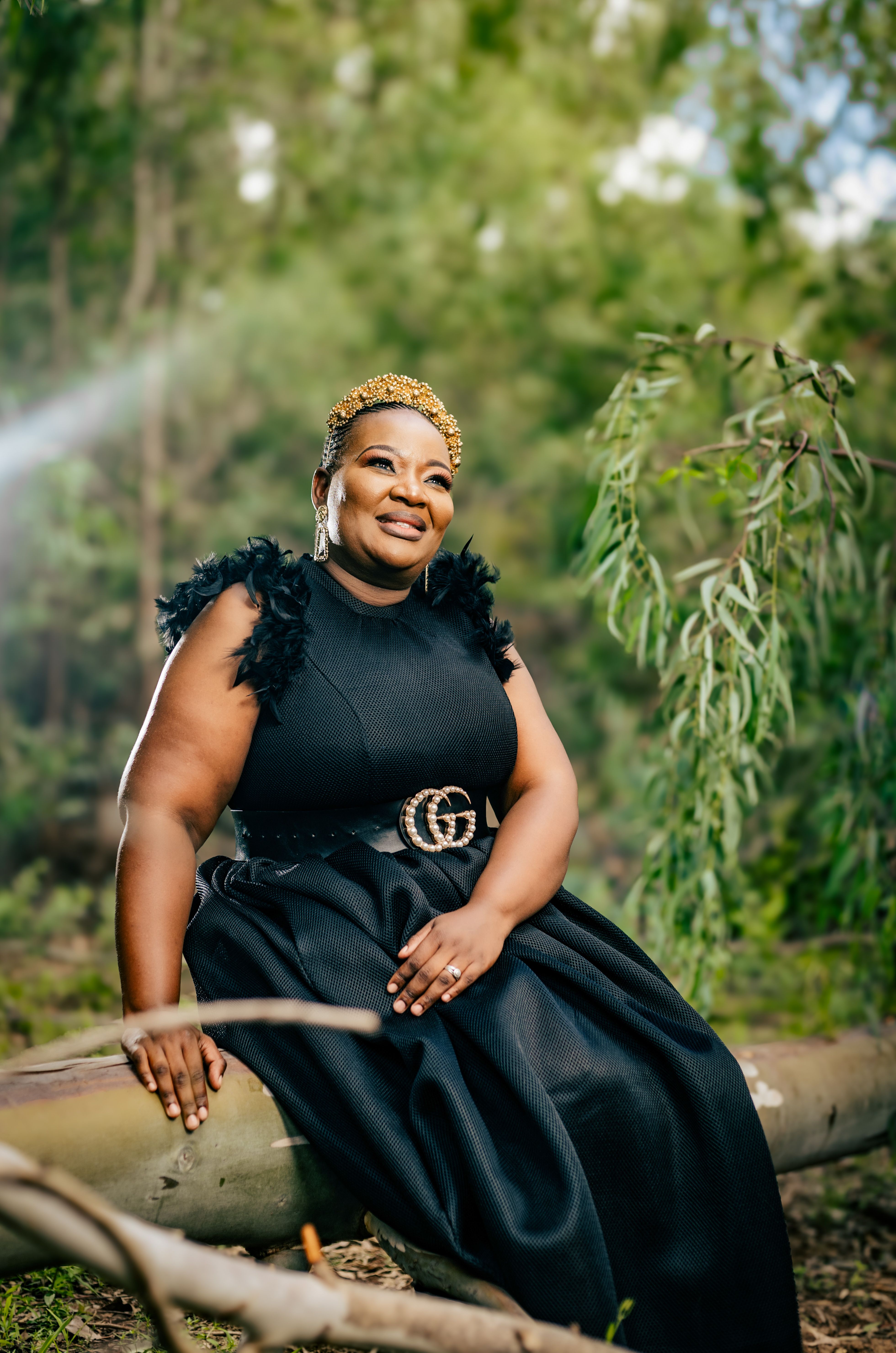 A woman in a black dress sits on a log in a lush forest, exuding elegance.