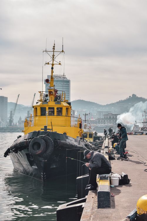 Free Men fishing at a harbor with a yellow tugboat and mountains in the background. Stock Photo