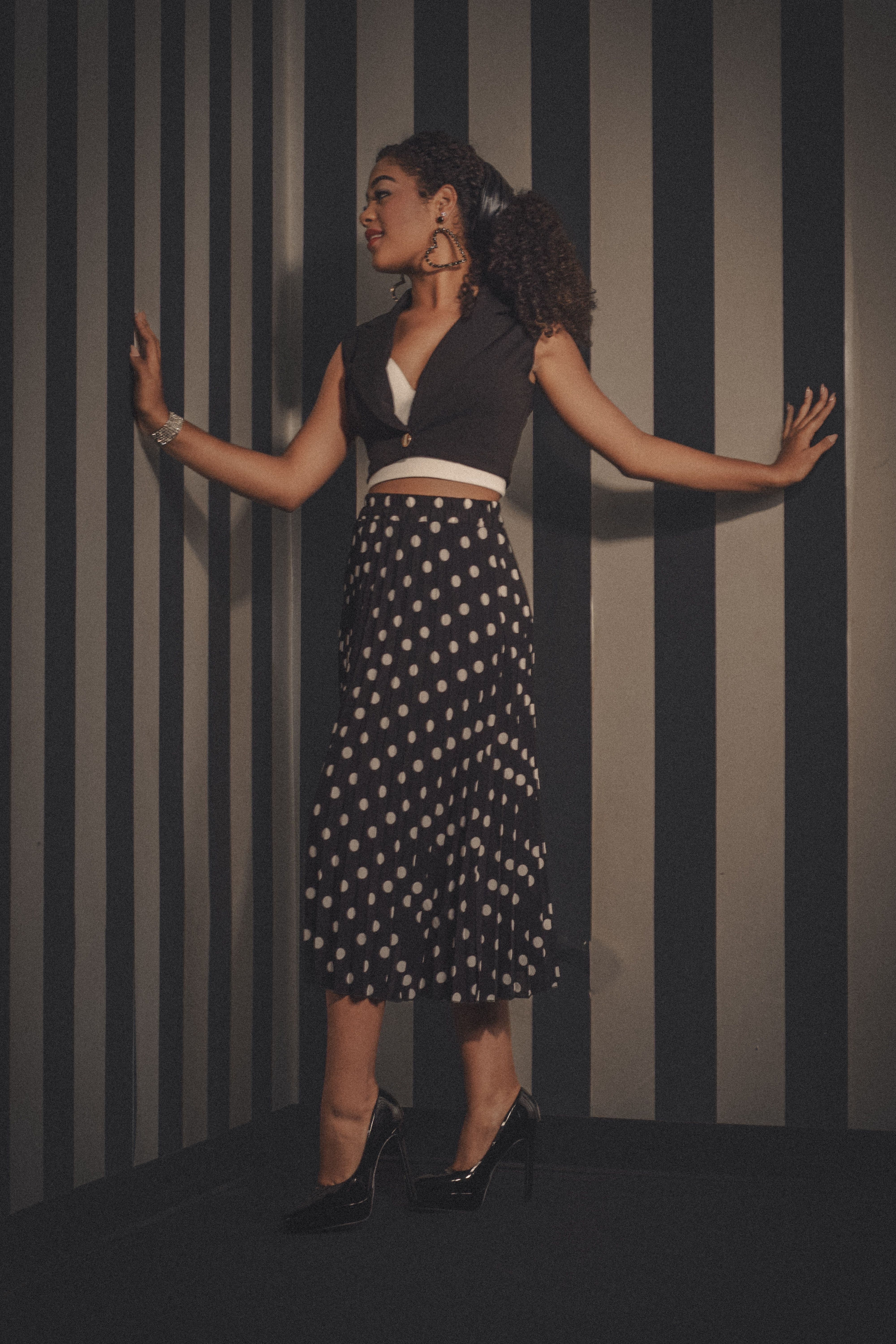 A woman elegantly posing in a polka dot dress against a striped wall indoors, showcasing modern fashion.