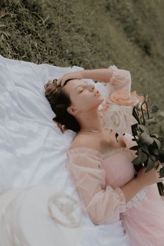 A serene woman in a pink dress lies on grass holding a bouquet of roses.