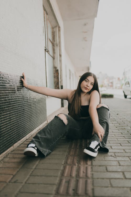 Young Woman in Ripped Jeans, Crop Top and Sneakers Sitting on the Sidewalk in City 