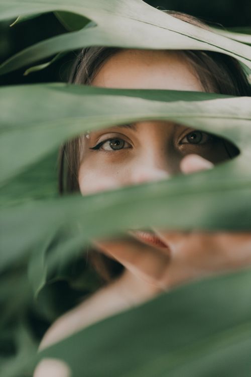 Free Mystical portrait of a woman peeking through lush tropical leaves, creating an enigmatic and serene mood. Stock Photo