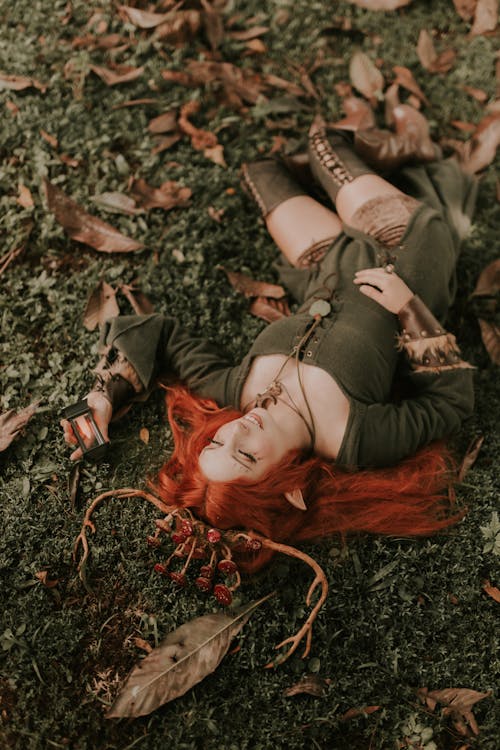 Woman with Ginger Hair Wearing a Headband with Antlers and Mushrooms Lying on the Ground in a Forest 