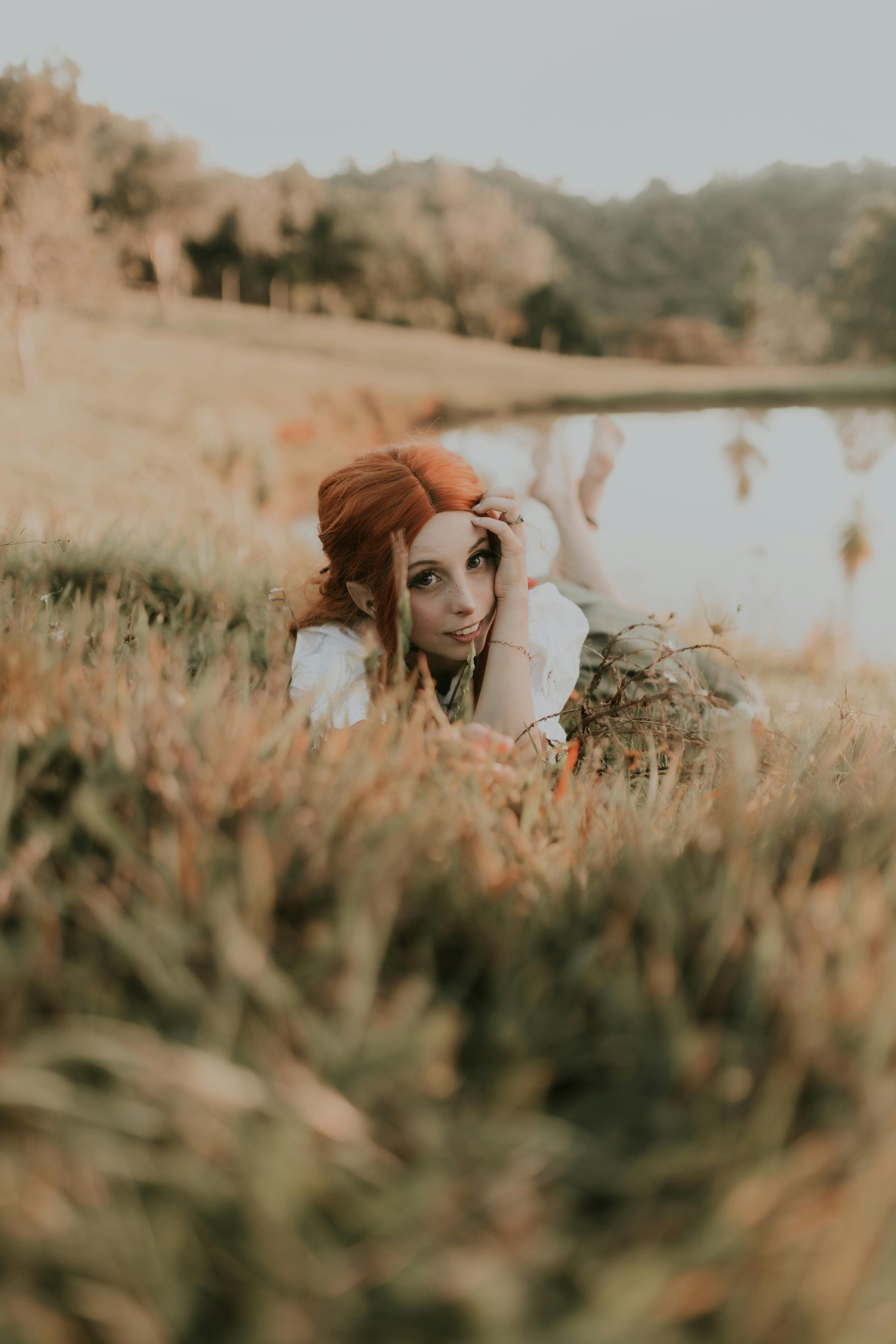 Whimsical redhead model lying in lush grass by a tranquil lake, exuding ethereal charm.