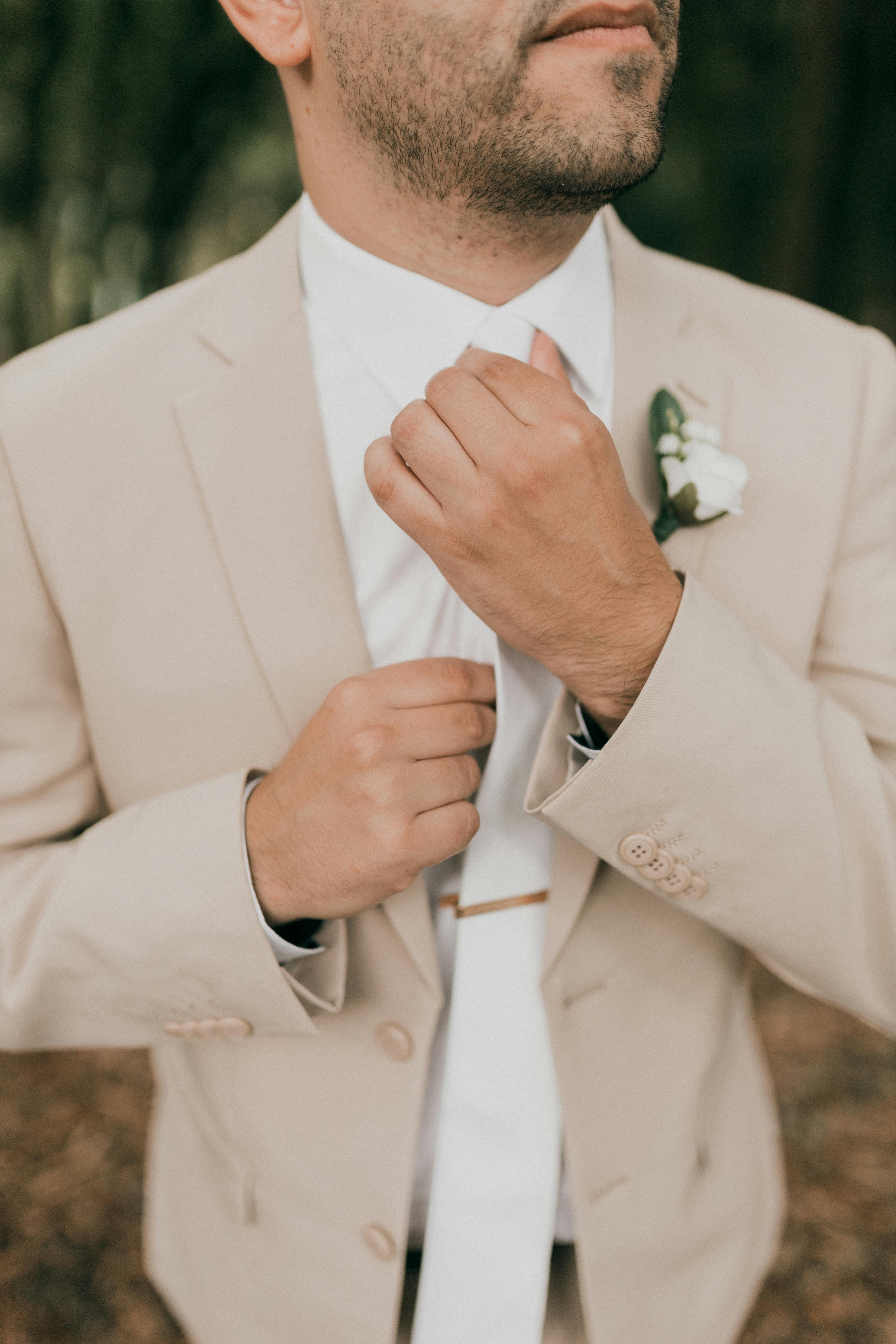 Close-up of a groom adjusting his tie in a stylish beige suit with a boutonniere.