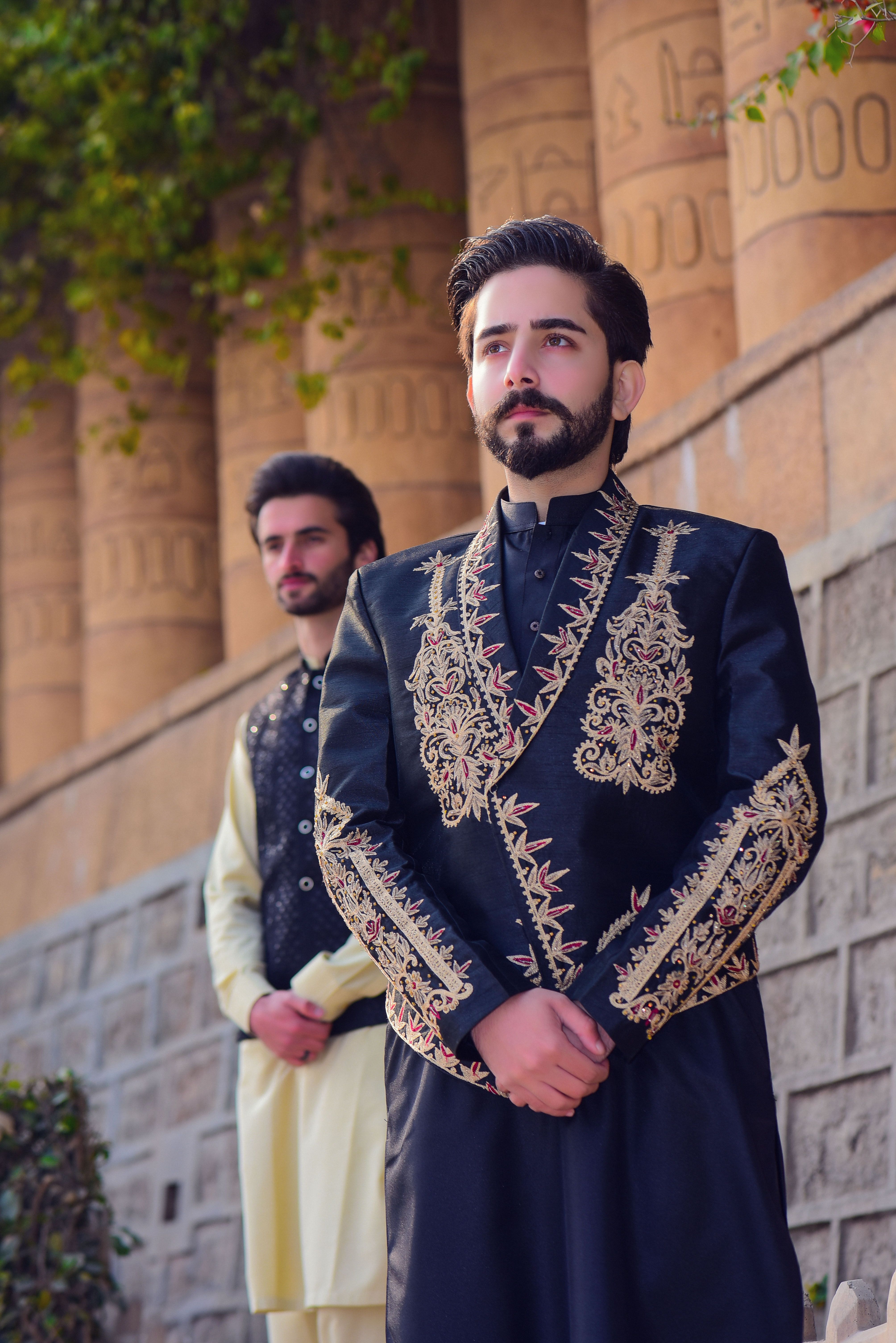 Two men in ornate traditional clothing pose outdoors, showcasing embroidery and fashion style.