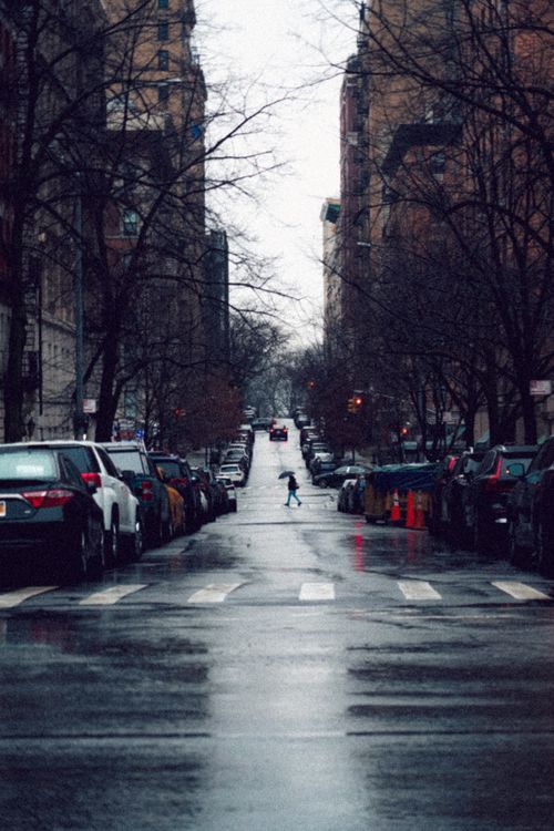 Free A rainy urban street scene with parked cars, a crosswalk, and a person crossing, creating a moody city atmosphere. Stock Photo