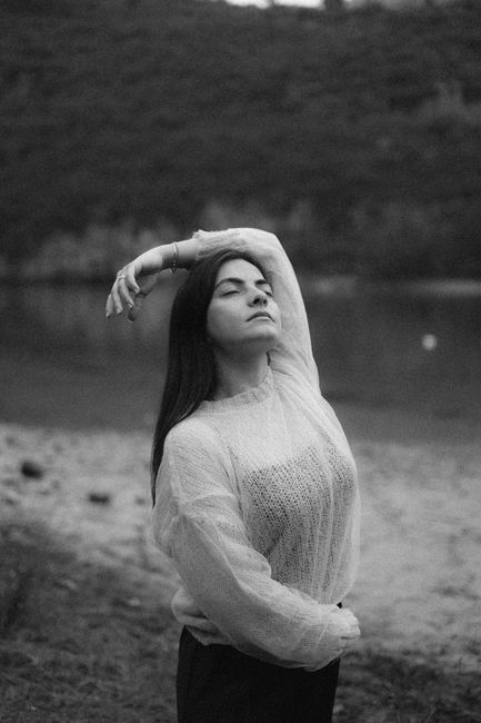 A woman with eyes closed, elegantly posing by a riverside in black and white.