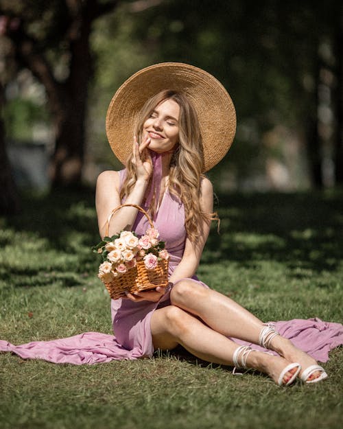 Elegant Woman in a Dress and a Sunhat Sitting on the Grass with a Basket of Flowers