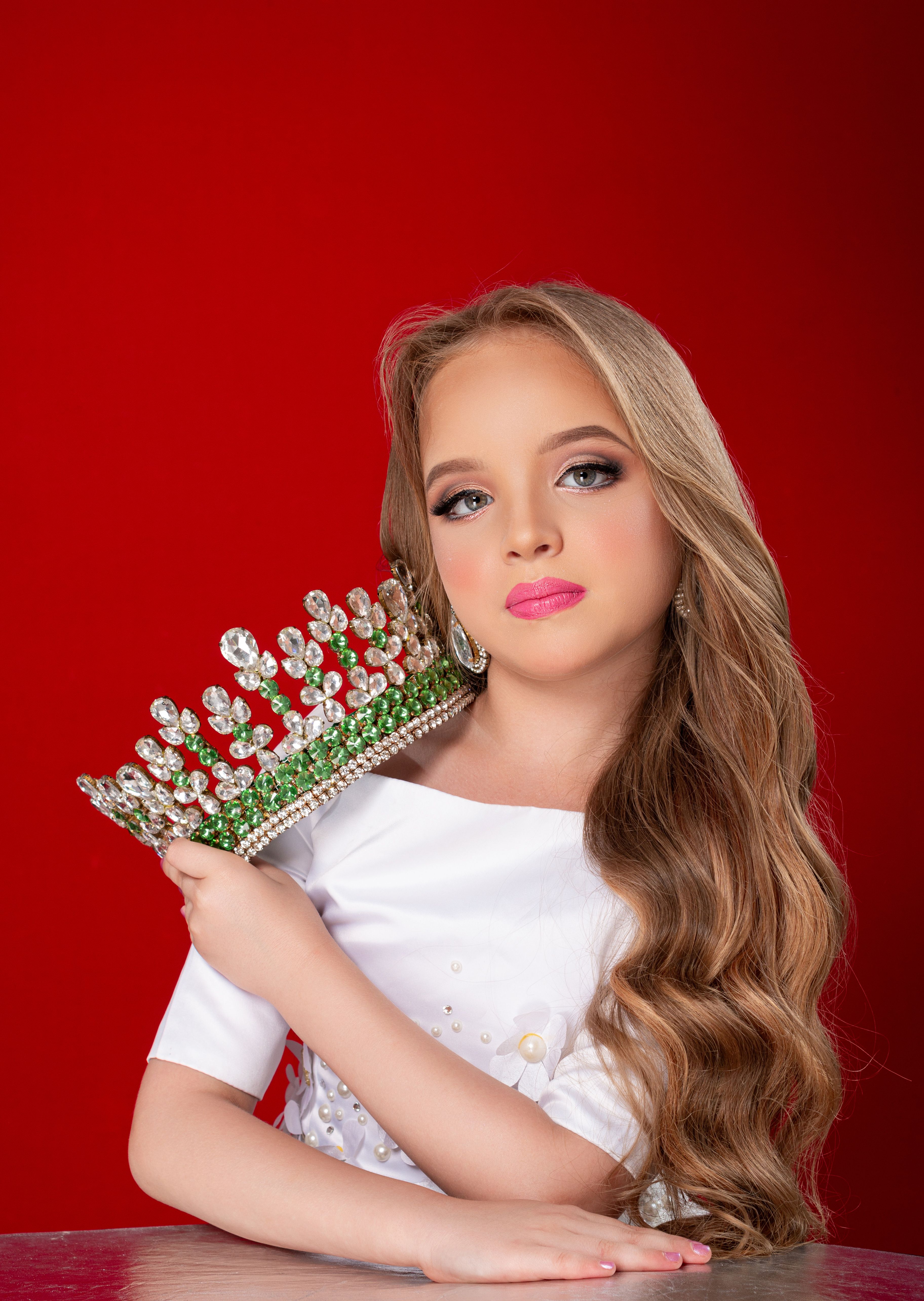 Fashion portrait of a young girl holding a jeweled crown in front of a vibrant red background.