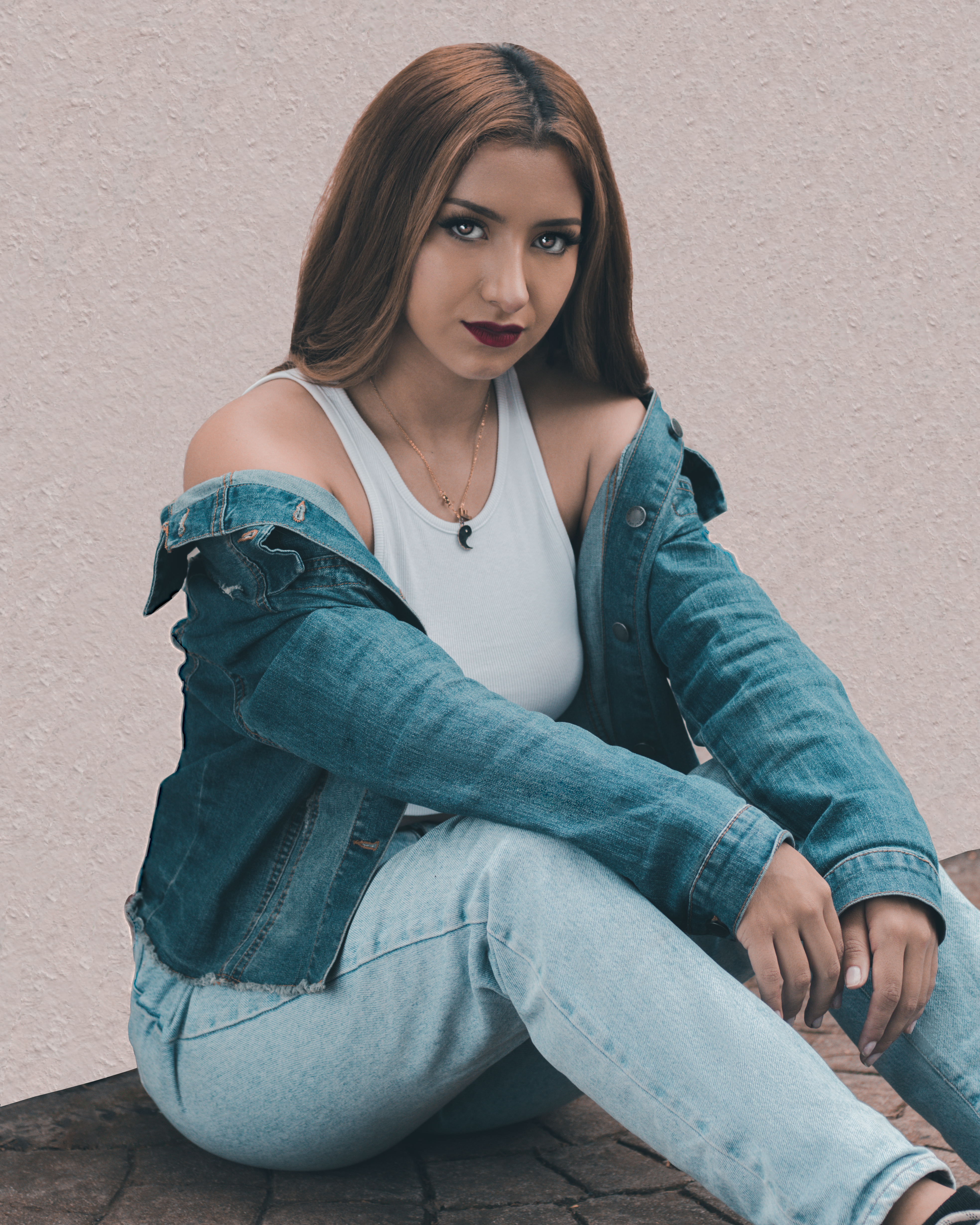 Portrait of a woman in a white tank top and denim jacket, sitting against a light background.