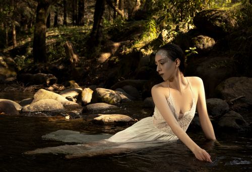 Free Fashionable woman in a white dress poses elegantly by a rocky stream in a serene forest setting. Stock Photo