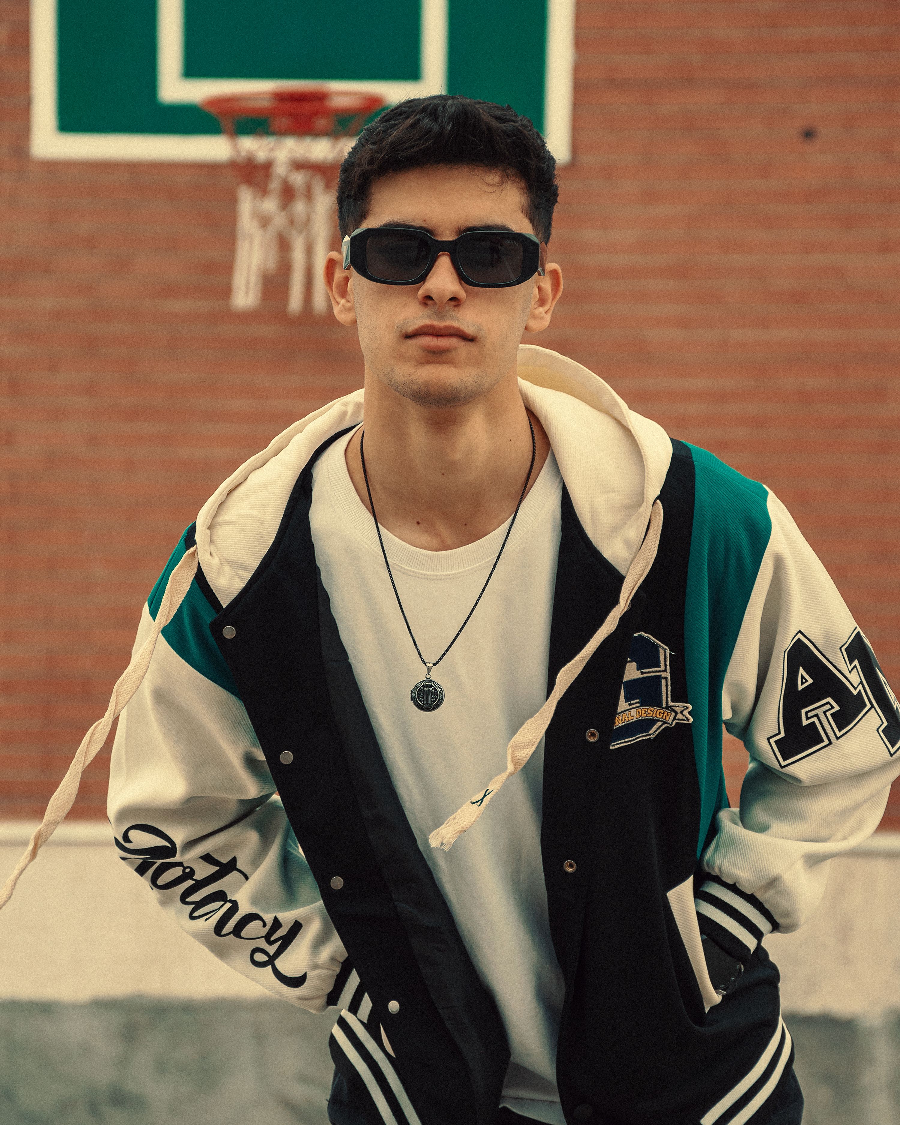 Young man wearing sunglasses and varsity jacket stands confidently outdoors near basketball hoop.