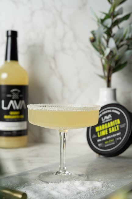 Two salt-rimmed margarita glasses filled with a pale gold cocktail, garnished with lime wedges, next to a small dish of coarse salt and a bottle of agave nectar.