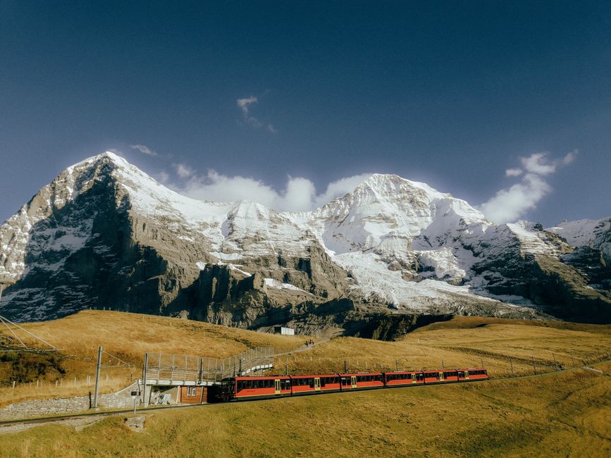 Captivating view of a red train traversing snowcapped Alps, under a clear blue sky.