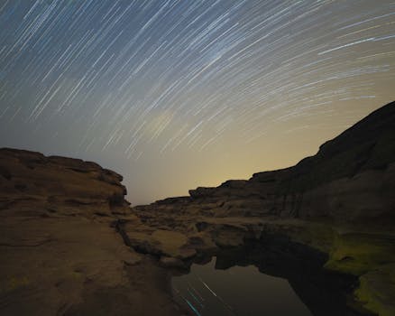 Dramatic night sky with star trails above a barren and eroded canyon landscape.