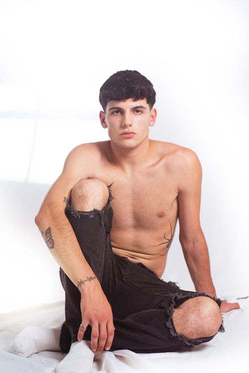 Free Confident young man sitting in a studio, wearing torn jeans, against a white background. Stock Photo