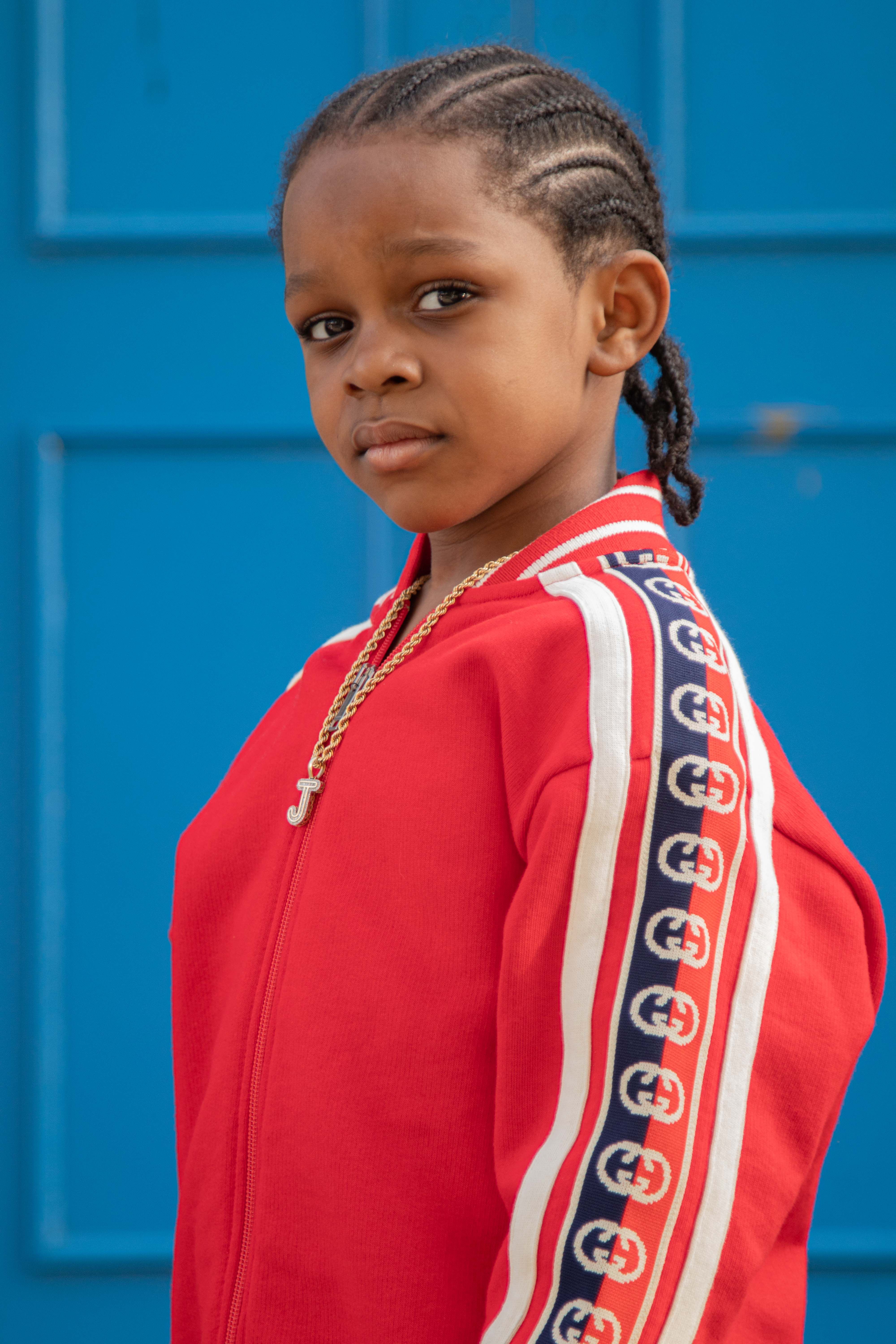 Portrait of a young child in a red jacket with braided hair, standing confidently against a vibrant blue background.