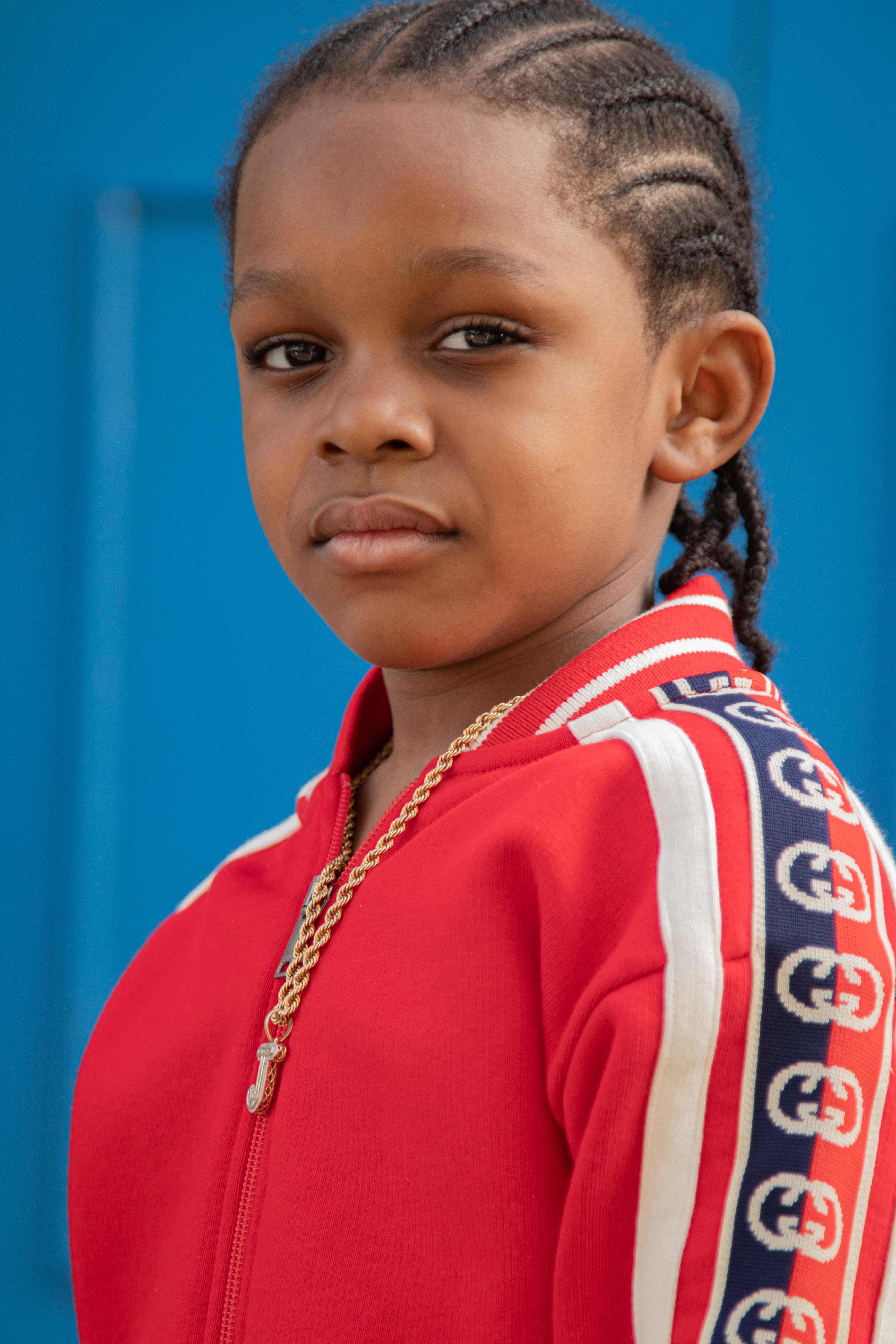 Young boy with braided hair in a red jacket, showcasing confidence against a blue background.
