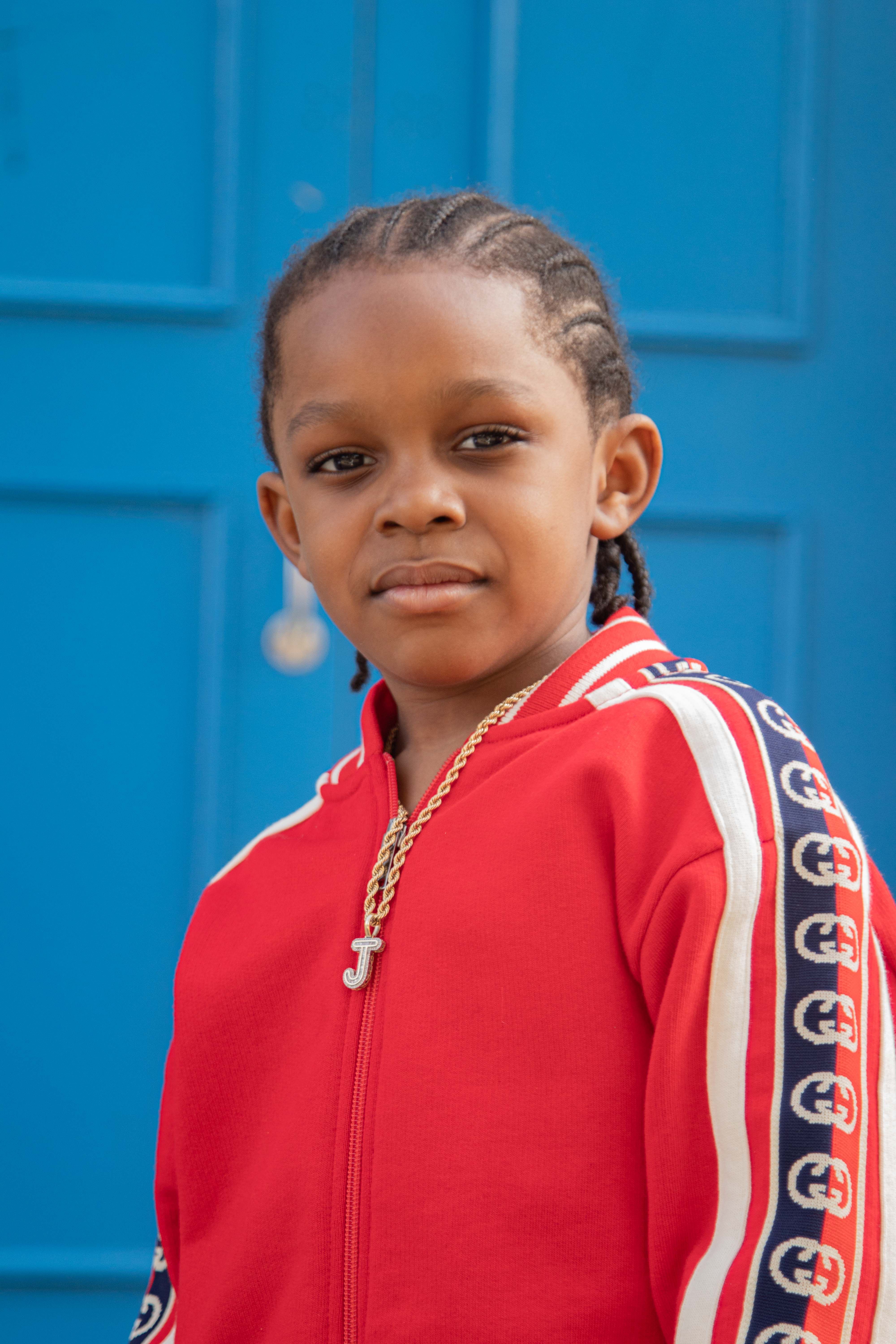 Portrait of a young boy with braided hair and jewelry in a red tracksuit standing in front of a blue door.