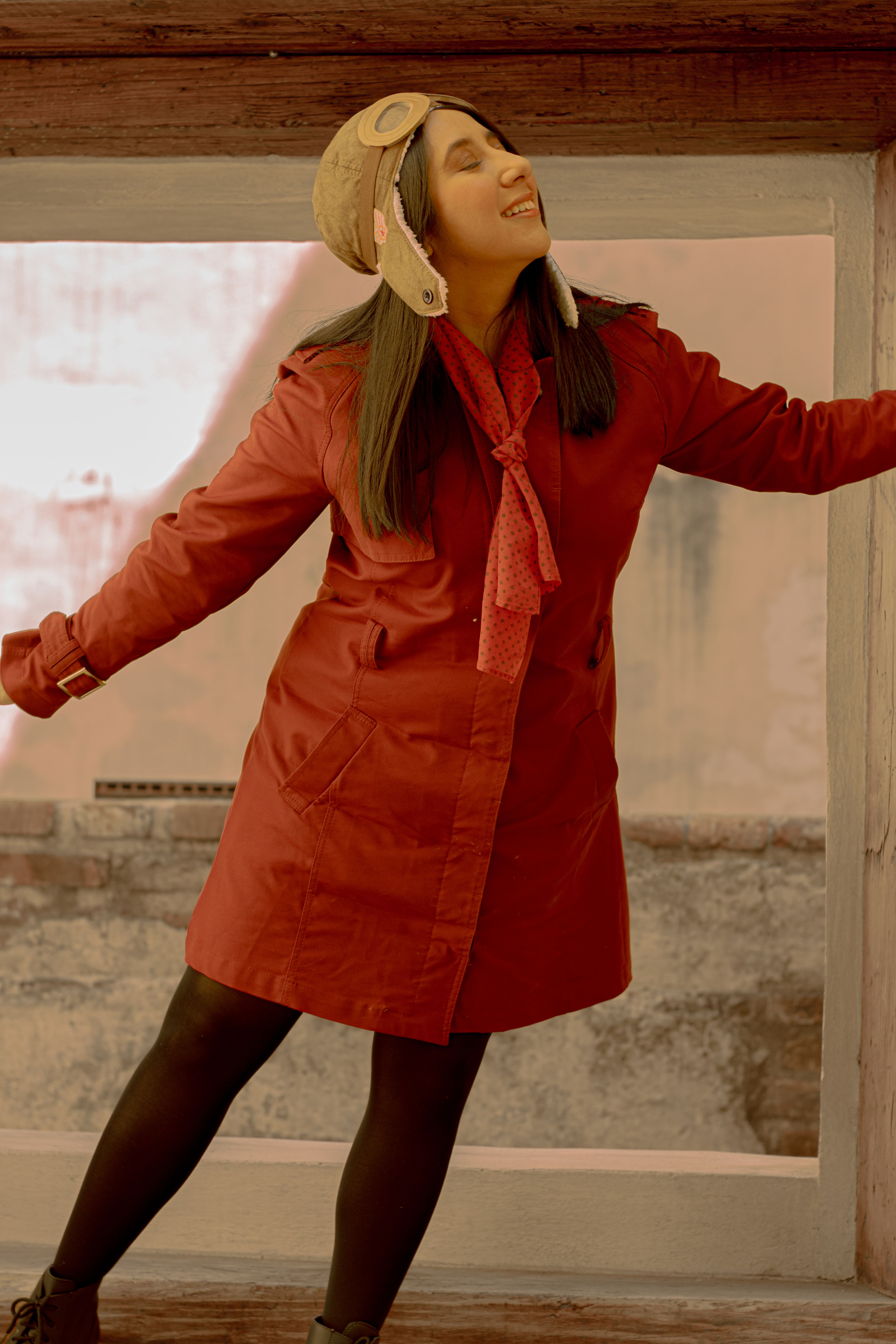 Smiling woman in a red coat and aviator hat posing playfully by a window indoors.