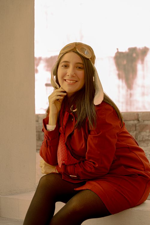 Free A young woman smiles warmly, dressed in a red coat and aviator hat, sitting indoors. Stock Photo