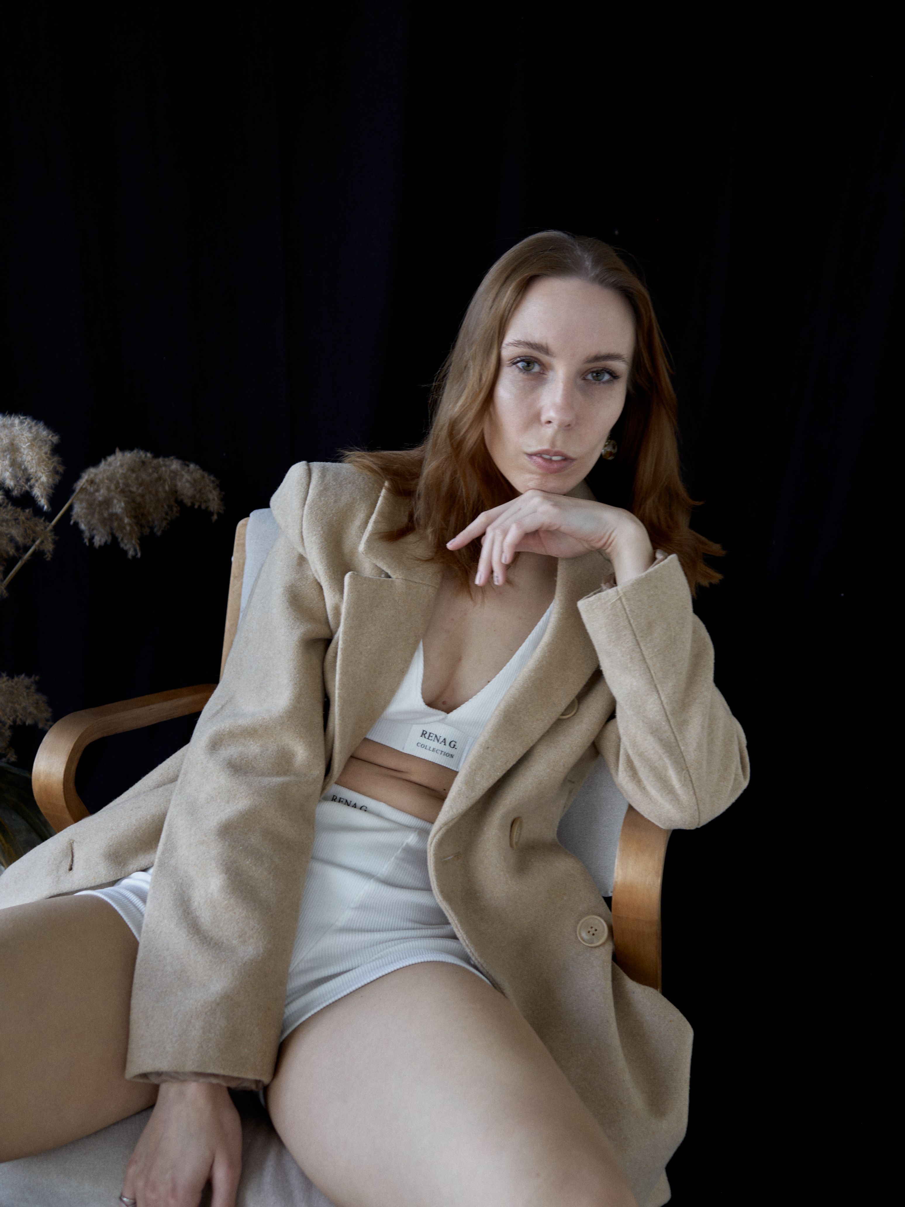 Fashion portrait of a woman in a beige coat, sitting confidently against a dark backdrop.