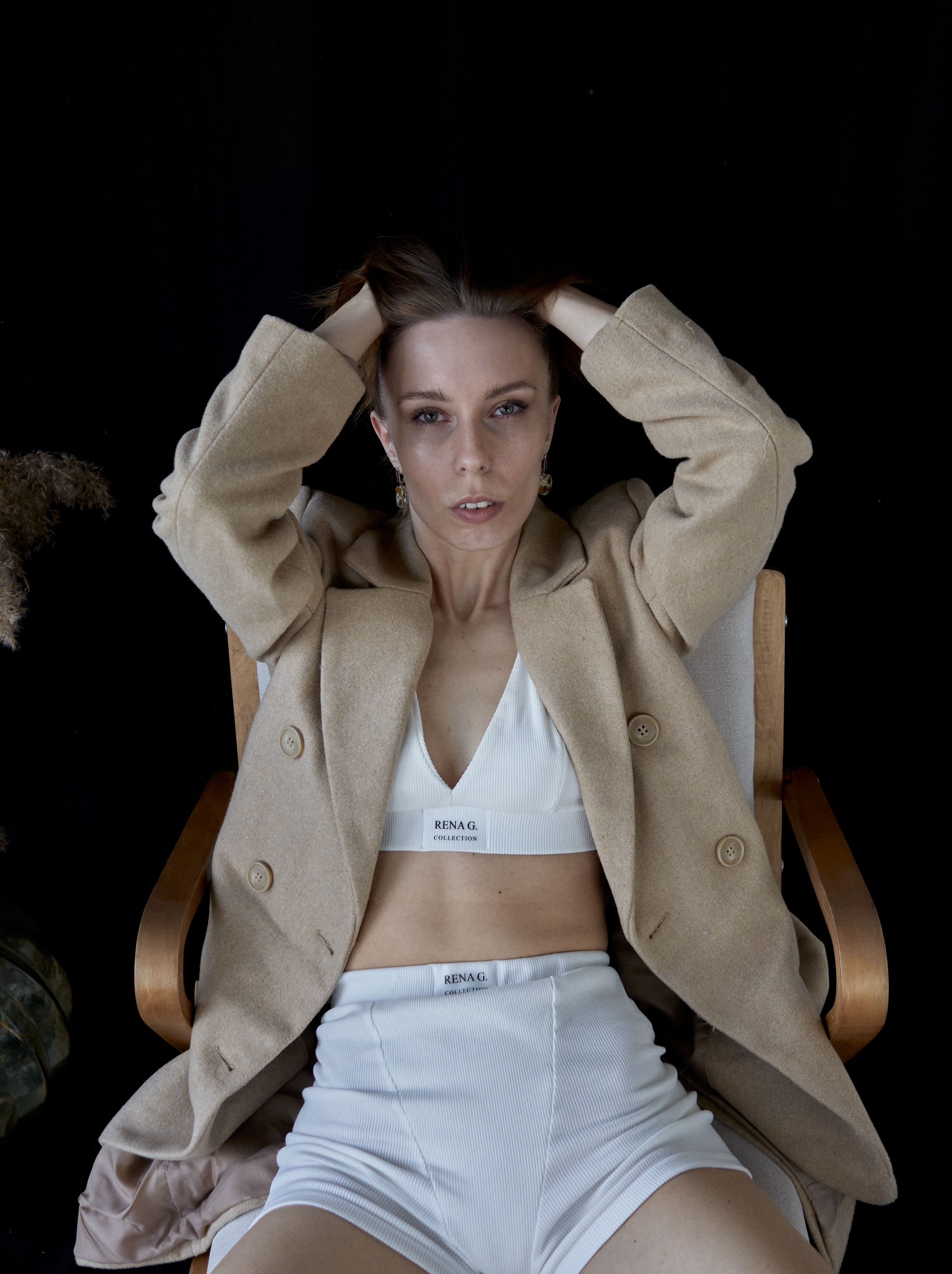 Stylish woman in beige suit and white lingerie posing against a black background in a studio setting.
