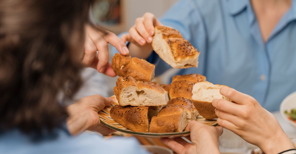 Family-style catering trays of baked ziti, garlic bread, and chicken parmigiana being passed hand-to-hand across a crowded, happy table