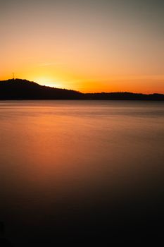 Tranquil coastal sunset view with smooth waters and silhouetted hills under an orange sky.