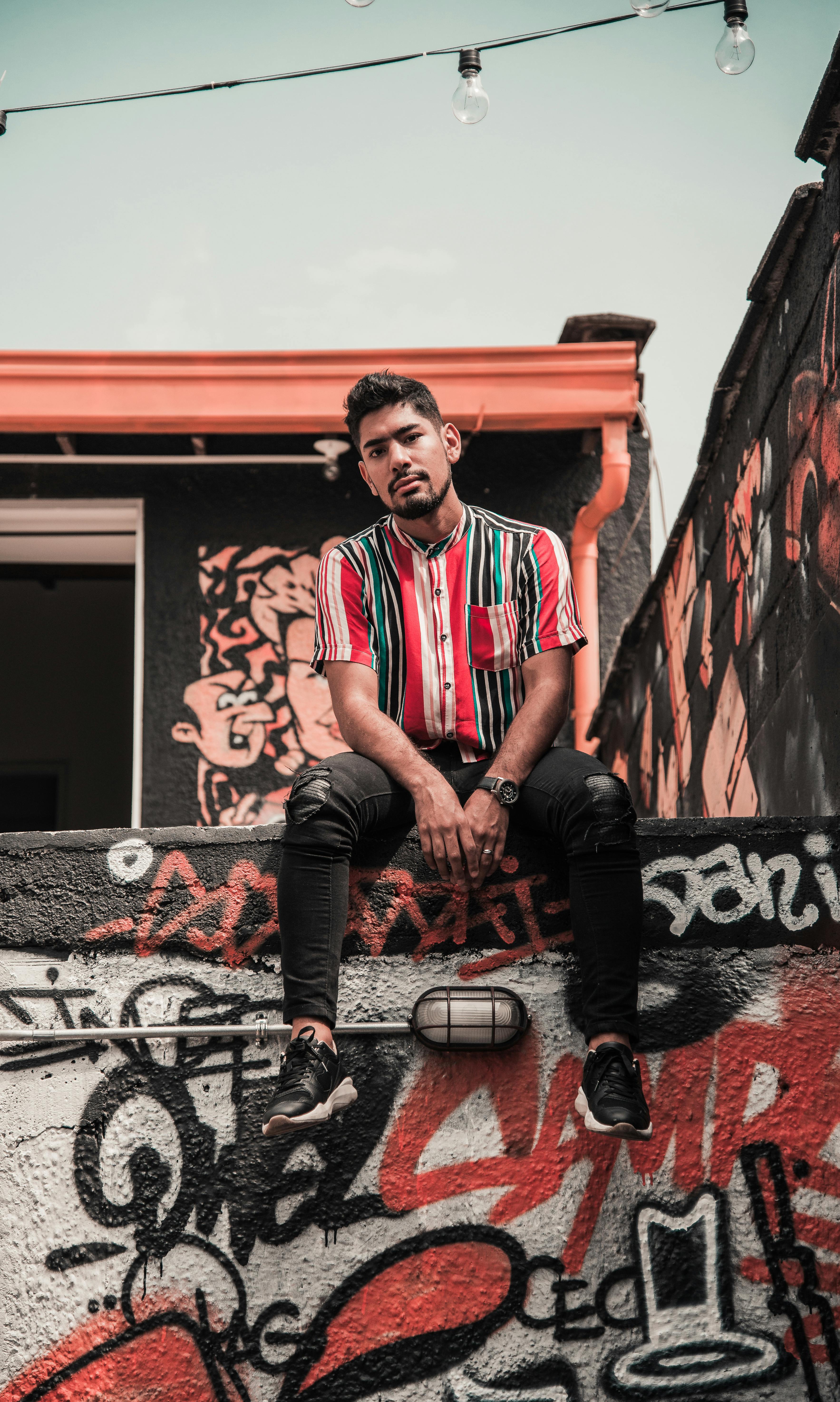 在哥伦比亚麦德林，一个留有面部毛发的年轻人穿着五颜六色的衬衫坐在涂鸦墙前