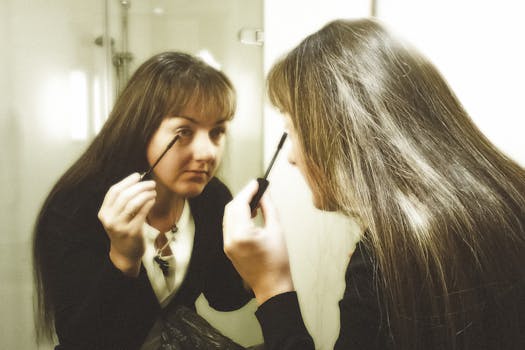 Brunette woman applying mascara in a bright bathroom mirror reflection.