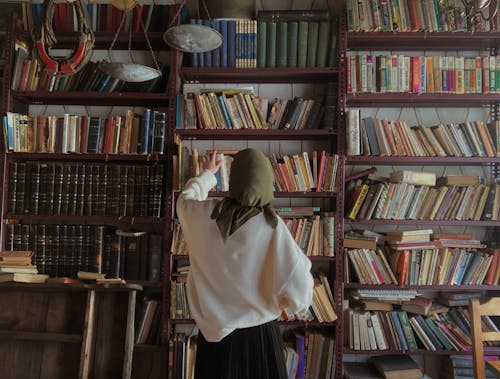 Woman in a Headscarf Taking a Book from a Shelf