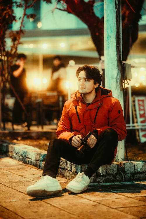 Young Man in a Jacket Sitting on a Curb on a Sidewalk in City at Night 