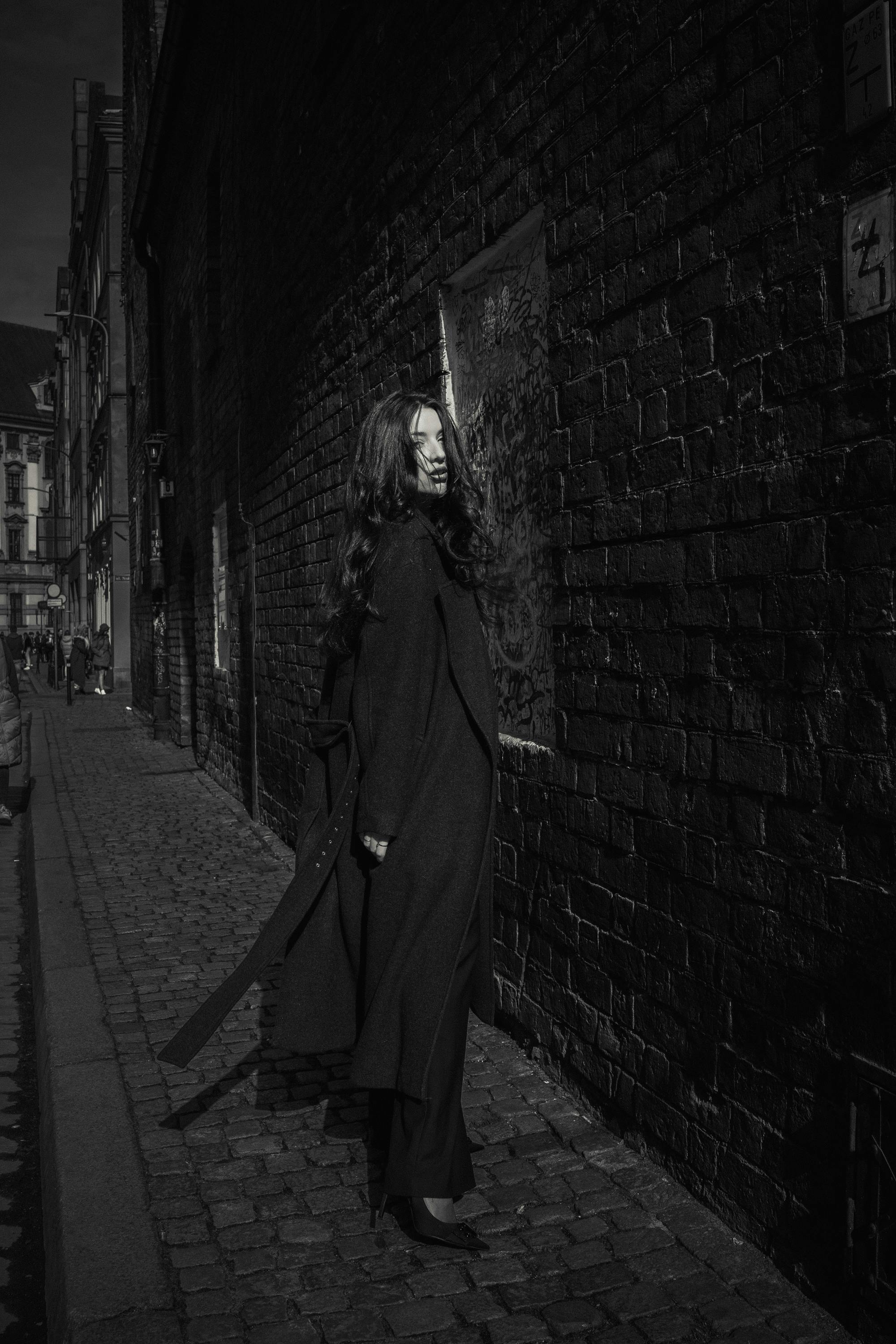 Black and white photo of a fashionable woman standing in an urban alley, highlighting long hair and coat.