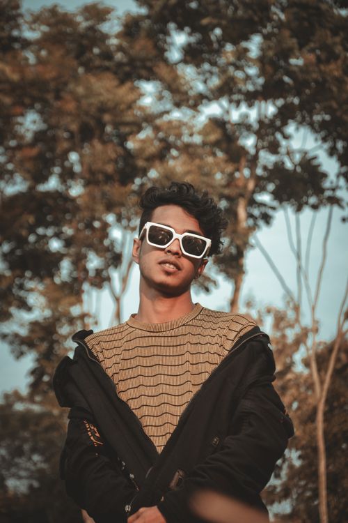 Free Stylish young man in sunglasses and jacket posing against trees under clear sky. Stock Photo