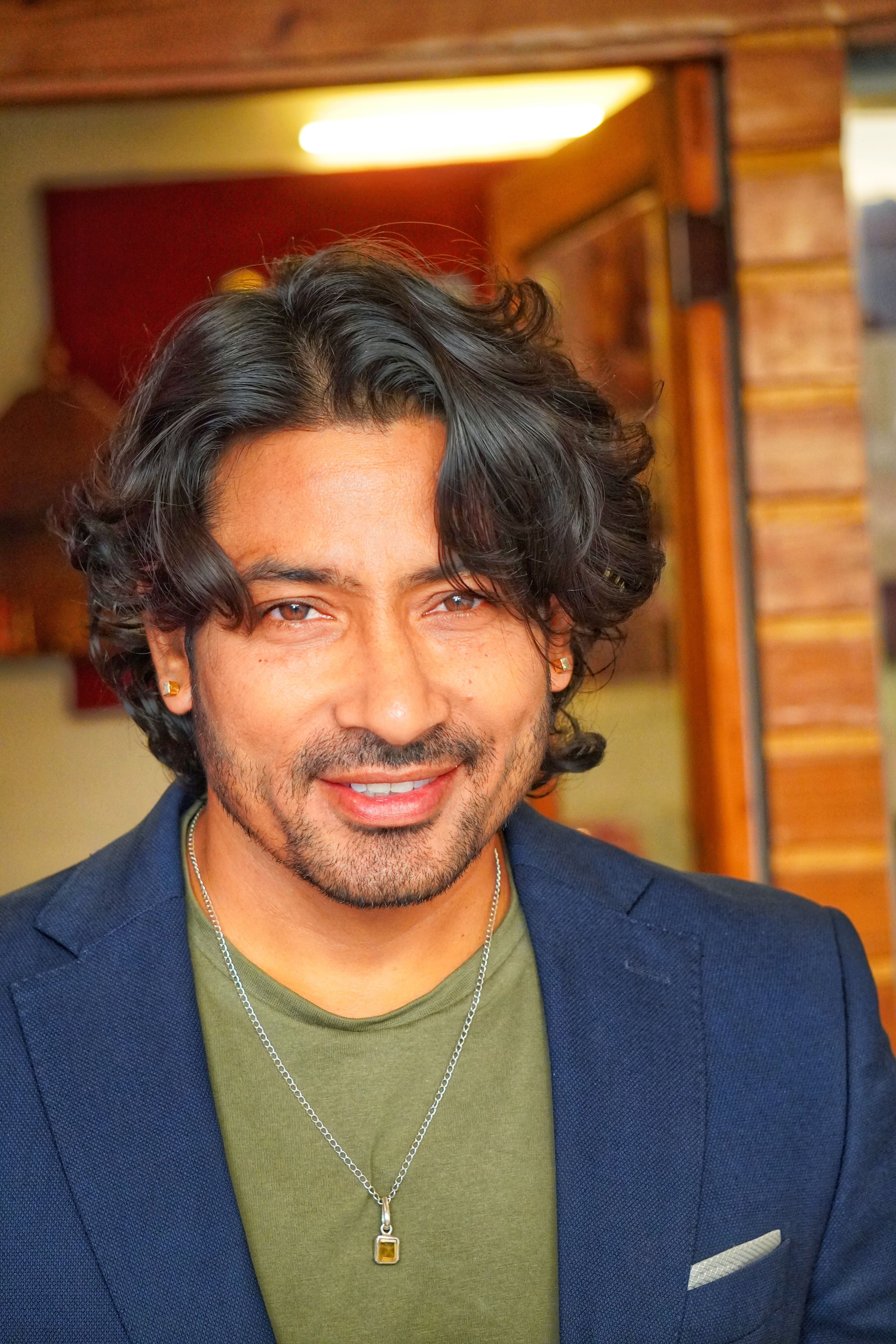A man with long hair smiling indoors, wearing a necklace and blazer.