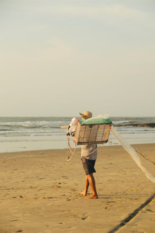 Free Fisherman carrying cage along the sandy shore at sunrise, serene ocean view. Stock Photo